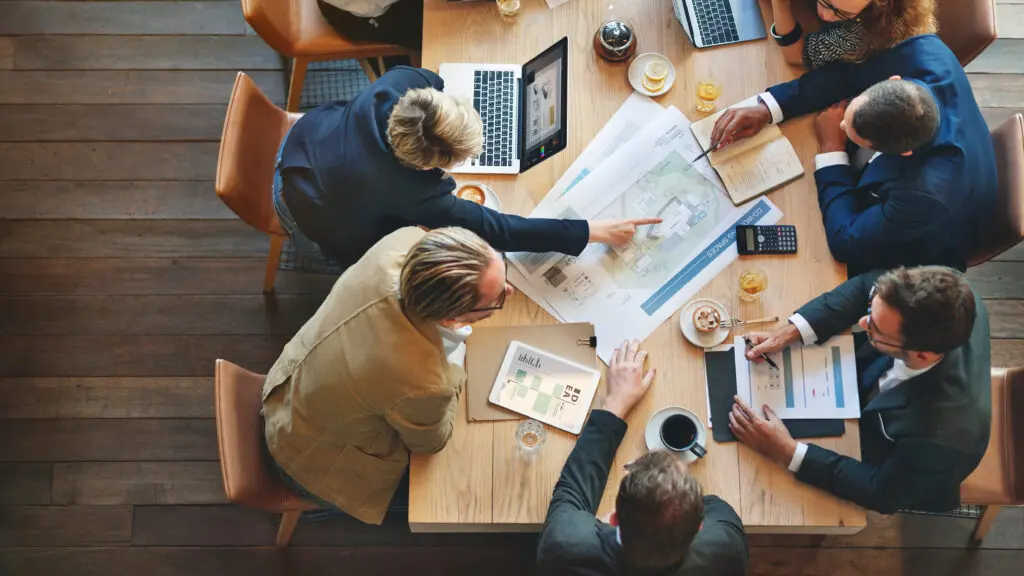 A group of business professionals sits around a wooden table in a modern office, collaborating on a project. One person points at an architectural blueprint, while others take notes and review documents. Laptops, coffee cups, and glasses of water are scattered across the table.