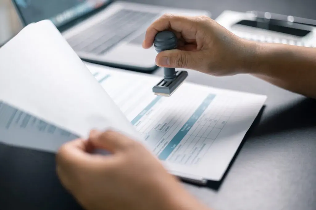 A close-up of a person's hands stamping an official document with a rubber stamp. The document contains tables and structured data, and a laptop is visible in the background on a dark desk. The scene suggests an administrative or legal approval process.